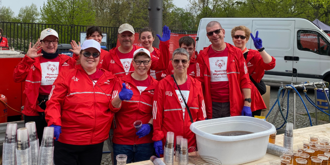 Ein inklusives Helfer*innen Team aus zehn Personen. Alle tagen rote T-Shirts und Jacken mit dem Special Olympics Deutschland Logo. Vor ihnen steht ein Tisch mit vielen Bechern und einer kleinen Wanne voll Tee.